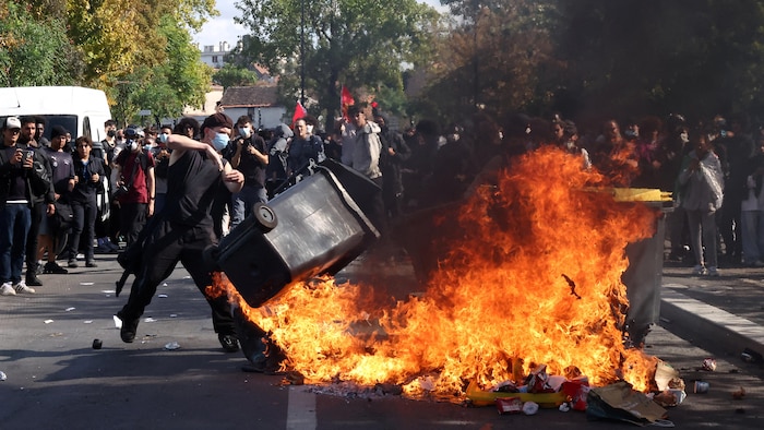 Un manifestant jette une poubelle dans un feu, devant d'autres manifestants.